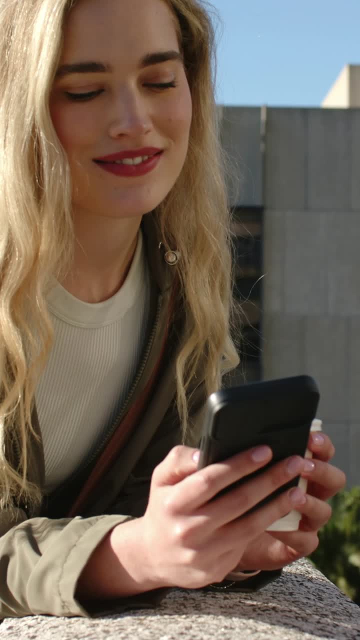 Video verticale: Una donna bionda, guardando in basso, legge un messaggio su un telefono nero mentre si appoggia a una ringhiera sul tetto.