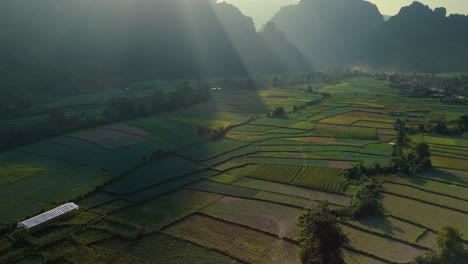 Hermosos-Rayos-De-Sol-Brillando-Sobre-Un-Fondo-Montañoso-Con-Un-Místico-Valle-Fértil-Y-Cultivos-De-Mosaico-En-El-Valle-De-Piedra-Caliza,-Vang-Vien,-Laos