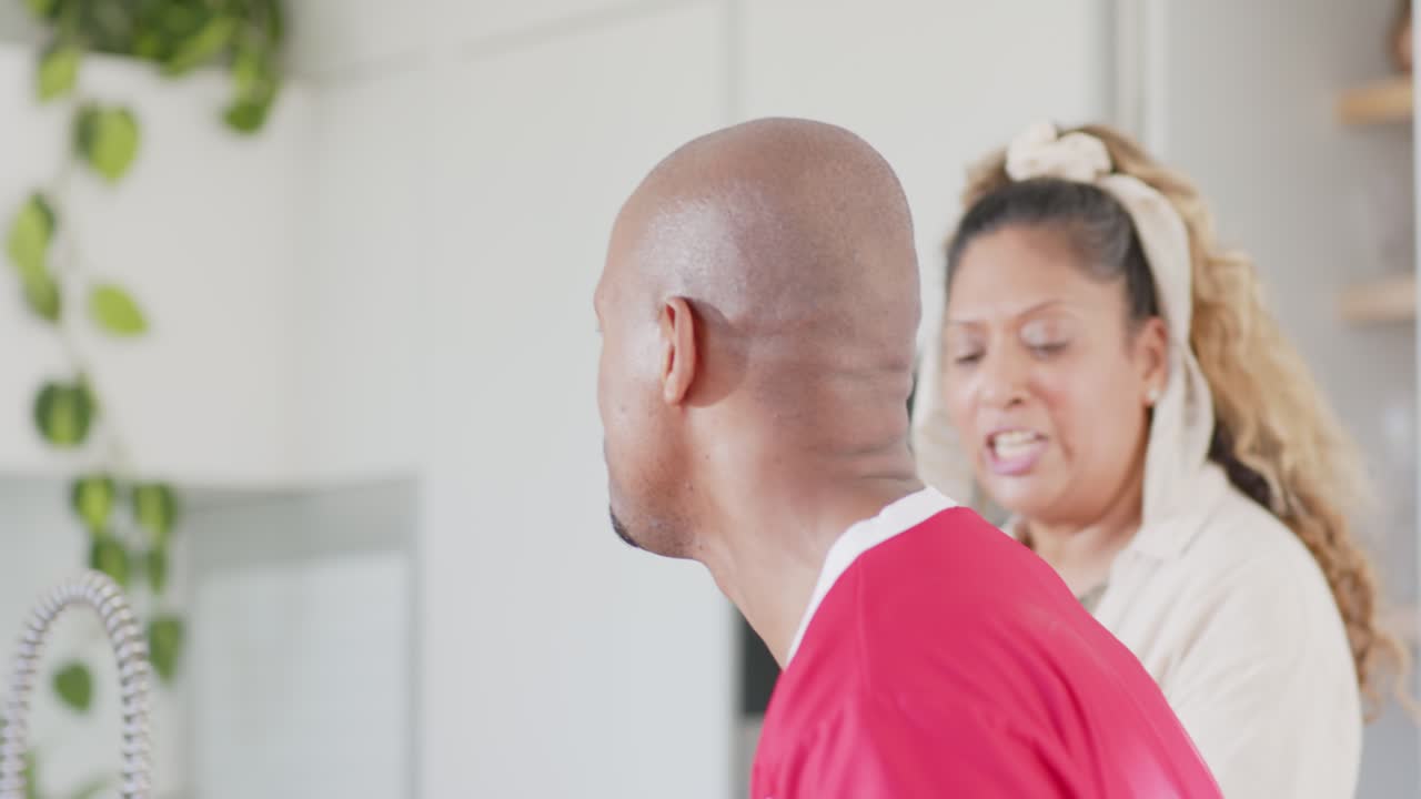 Diverse group seeing red jersey man holding soccer ball, replying to woman speaking in kitchen
