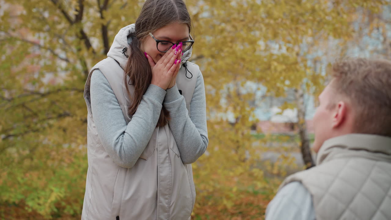 Close up of boyfriend kneeling to propose to smiling girlfriend with painted nails who covers her mouth in excitement and nods in agreement surrounded by golden autumn leaves and residential building
