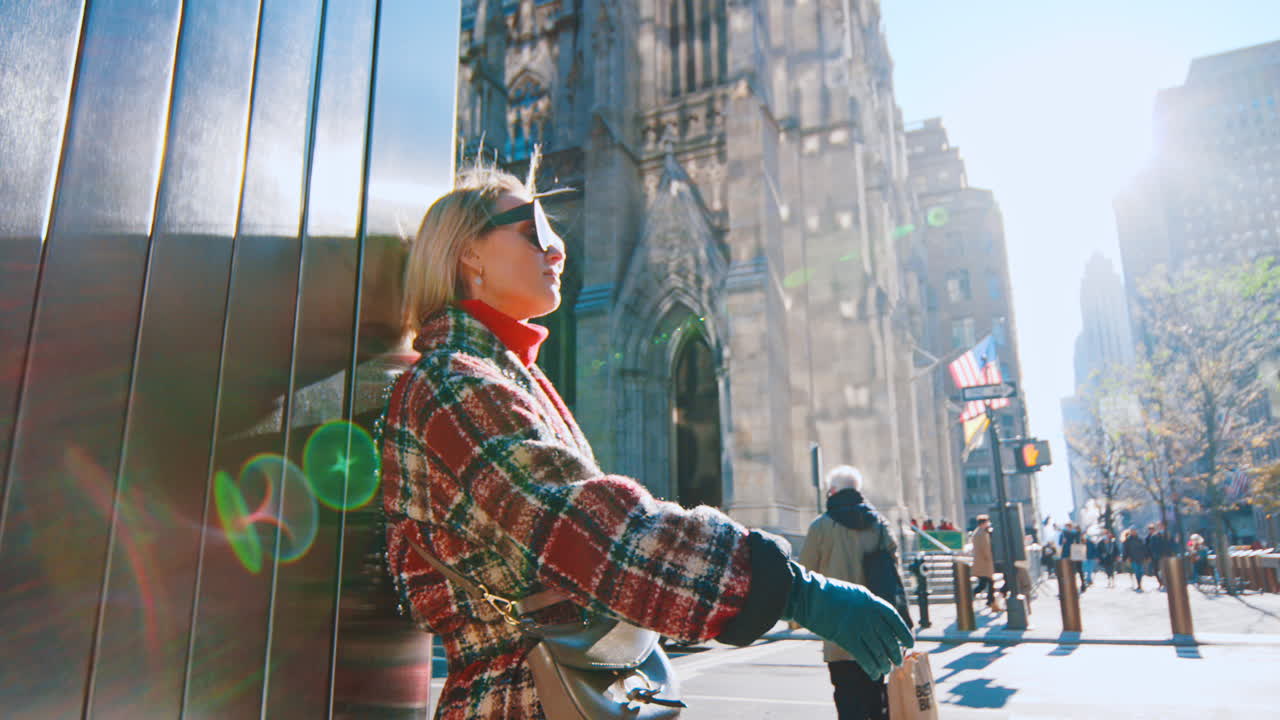 Nueva York, EE. UU., 25 de octubre de 2025: Mujer con los brazos cruzados apoyada en un pilar en Manhattan. Vista lateral de una mujer rubia con abrigo a cuadros y gafas de sol apoyada en un edificio en Nueva York.
