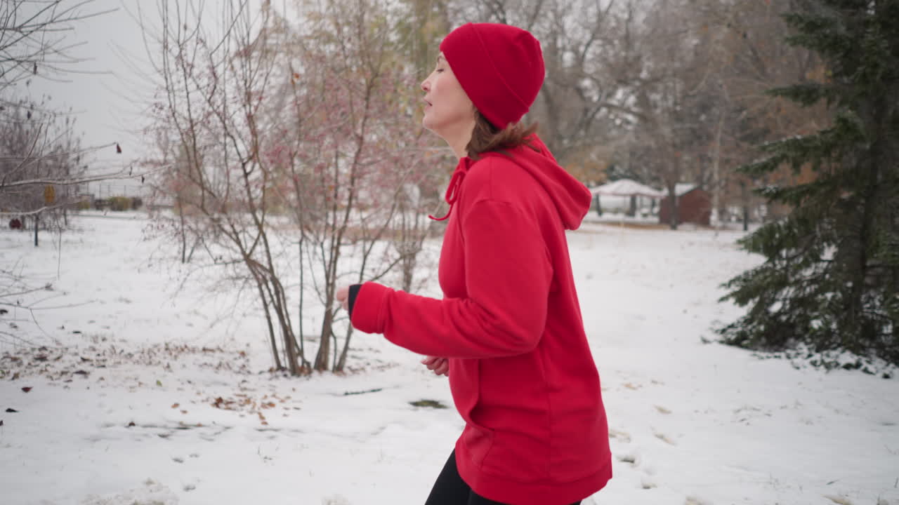 vista lateral de una mujer deportiva con gorra roja corriendo al aire libre durante el invierno rodeada de árboles cubiertos de nieve, pinos de hoja perenne y atmósfera brumosa con un edificio en el fondo lejano