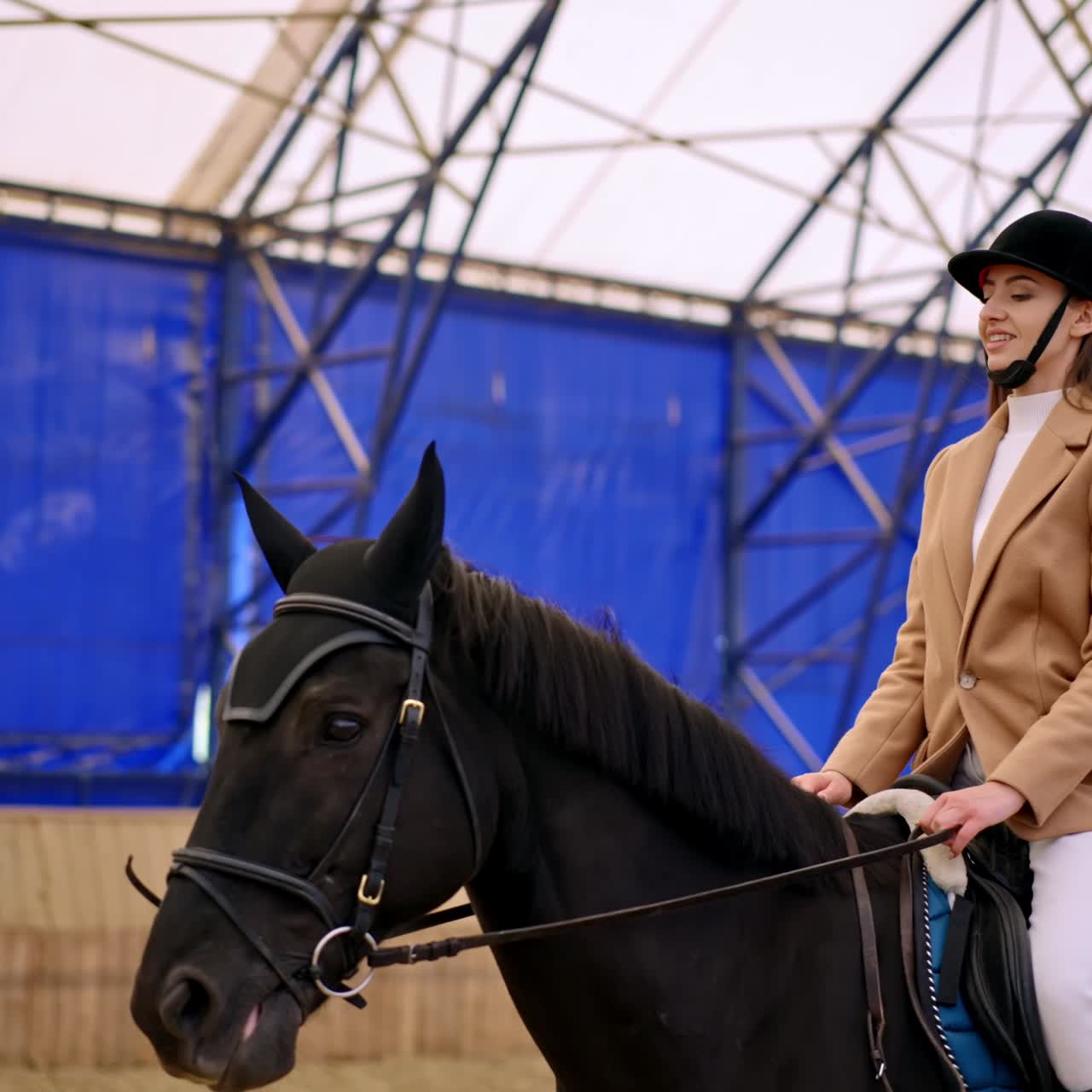 Caucasian woman in black jockey hat practicing horse riding. Lady takes riding lessons on a beautiful black horse. Manege at backdrop in blur