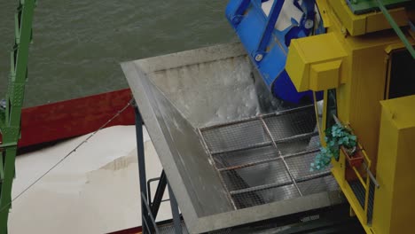 a crane unloads a ship filled with fertilizer at a port and drops it into a funnel