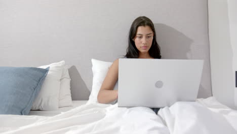 focused biracial teenage girl sitting in bed in the morning using laptop, copy space, slow motion