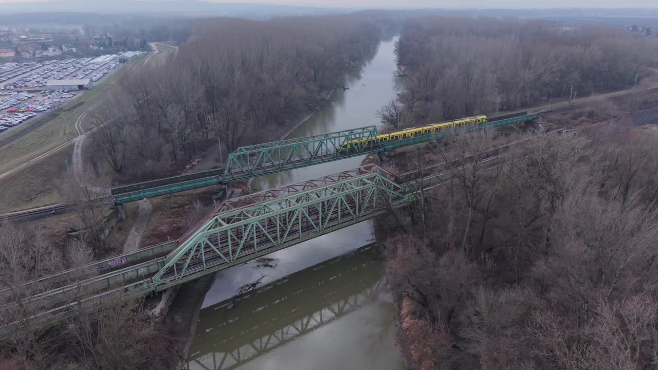Aerial view of a yellow passenger train crossing a steel railway bridge over a calm river surrounded by leafless forest