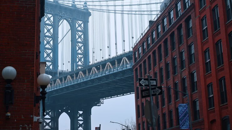 Steel suspension bridge seen between red brick buildings in urban setting. View of Manhattan Bridge tower framed by industrial red brick buildings in Dumbo district during foggy weather