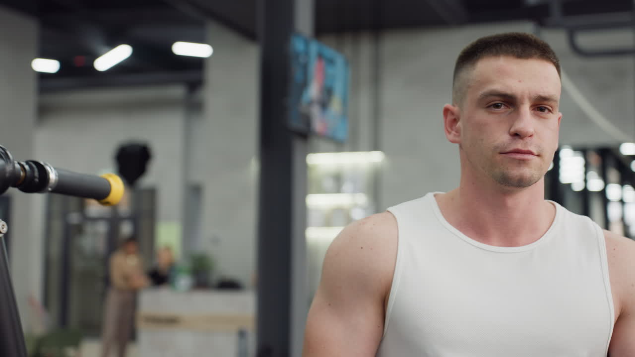 Portrait view of well bodied man walking through gym hallway past people working out on weight machines and cardio equipment under bright LED lights and industrial decor with small television on wall