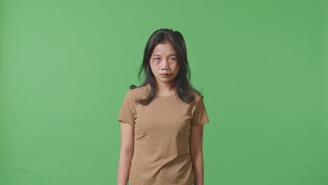 young asian woman victim of violence with bruise on face being offended looking at the camera on green screen background in the studio