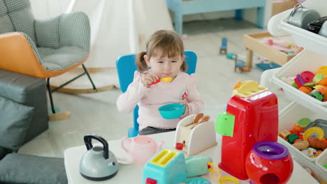 Lustiges-Kleines-Mädchen,-Das-So-Tut,-Als-Würde-Es-Mit-Einem-Spielzeuglöffel-Aus-Einer-Leeren-Plastikschüssel-In-Der-Küche-Essen-Und-Am-Tisch-Im-Spielzimmer-Sitzen