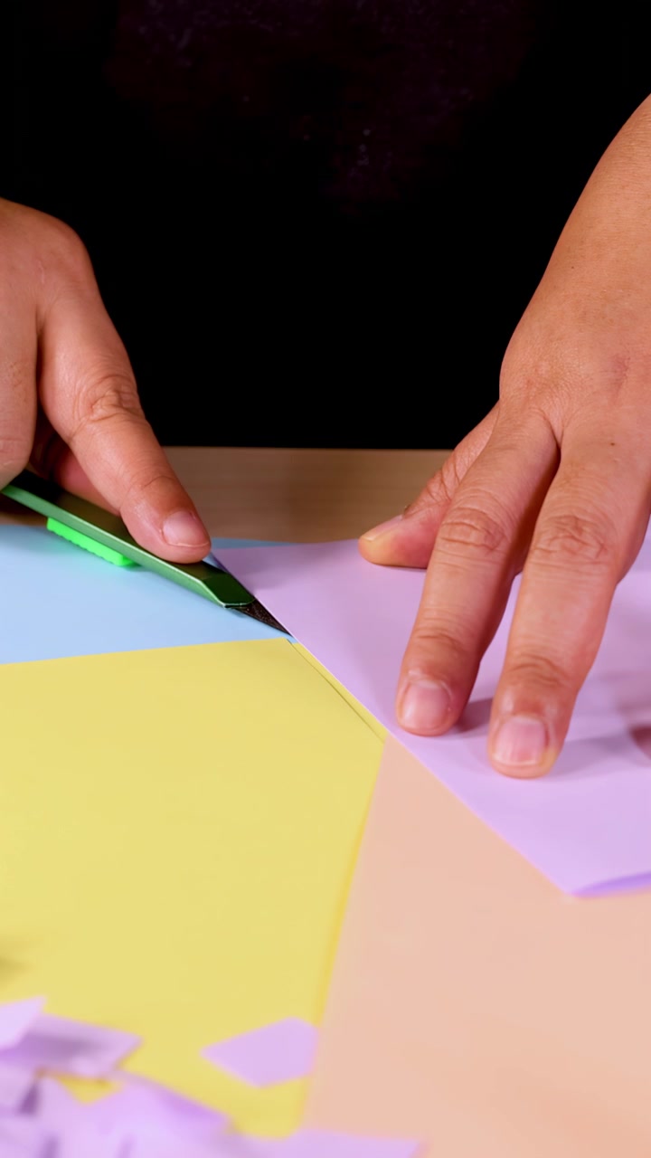 Hands use green craft knife to cut pastel paper on desk under bright, even lighting