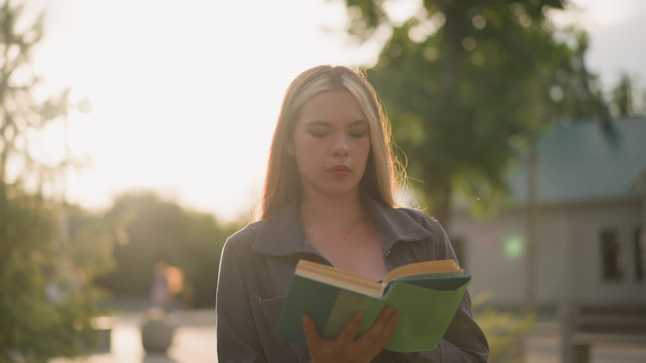 Lady in grey clothing walking on grassy field reading a book as sunlight creates bright flashes around her, with blurred view of trees and people in the background