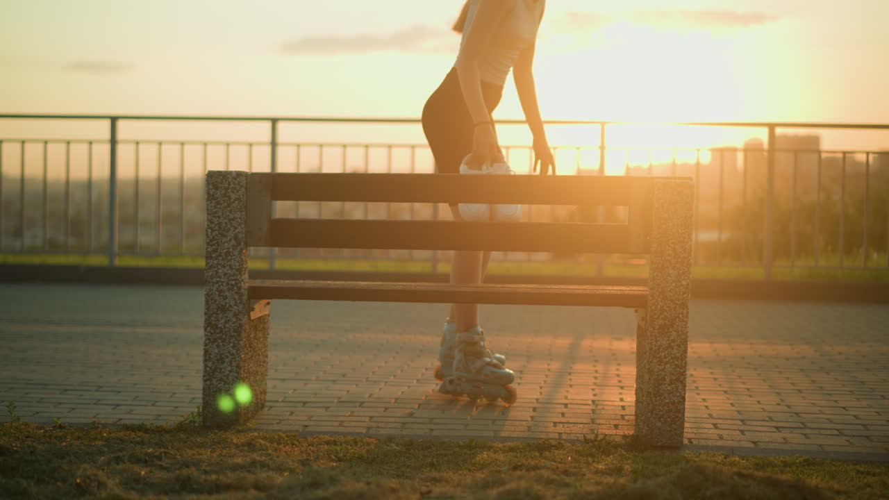 야외 벤치를 향해 롤러 스케이팅하는 젊은 여성은 저녁 해가 지는 동안 손에  스니커를 들고, 빛은 보행길에 긴 그림자와 함께 따뜻한 빛을 만니다.