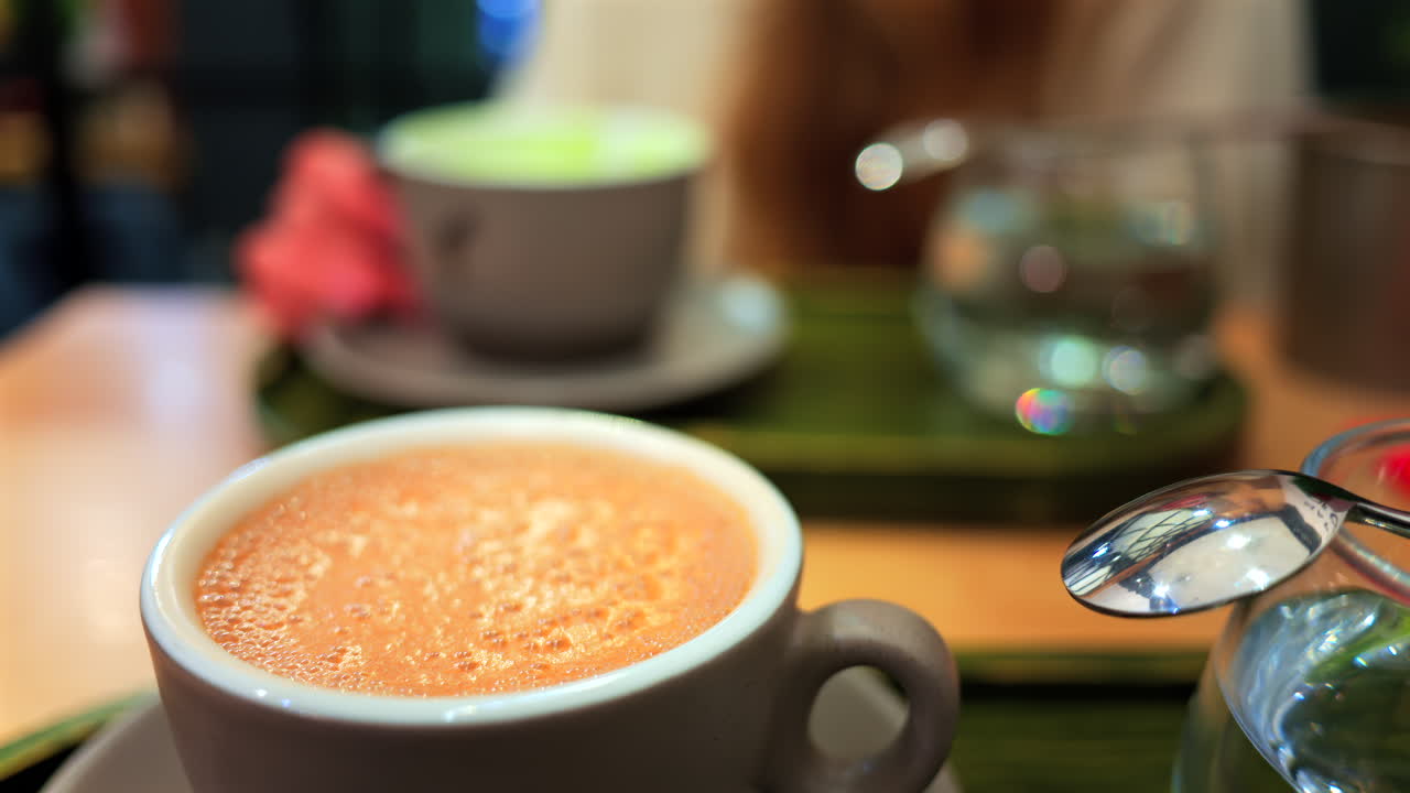 Close up of a cappuccino on a table at a cafe with a blurred view of a woman drinking a matcha latte on the background