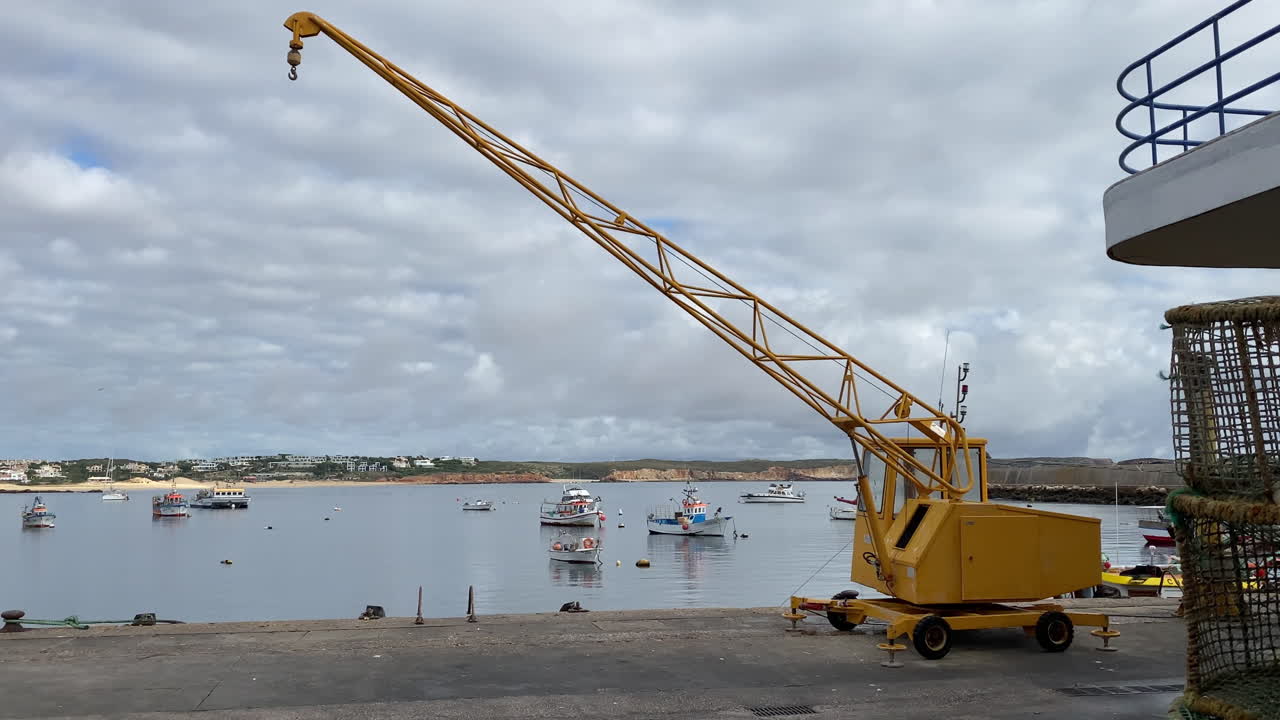 a Yellow harbor crane stands beside the calm fishing port of Sagres in the Algarve, with small boats floating on still water under a dramatic cloudy sky