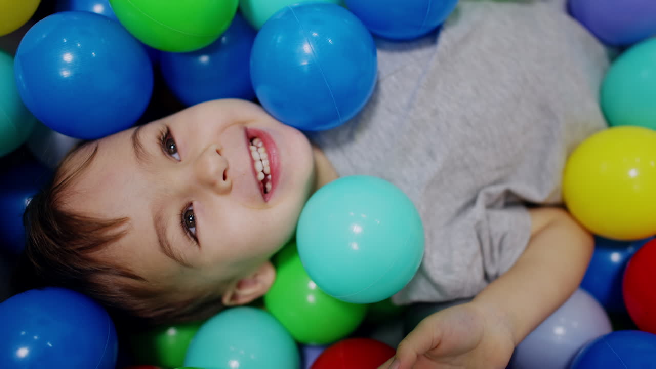 Happy Caucasian kid lies among the colorful balls. Baby boy plays in the dry pool. Close up.