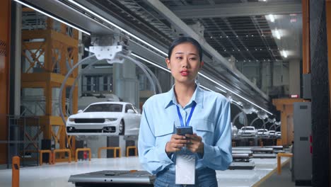trabajadora profesional asiática de pie con su smartphone en una fábrica de automóviles ev, instalación industrial de alta tecnología, observa mirando a su alrededor antes de concentrarse con su smartphone