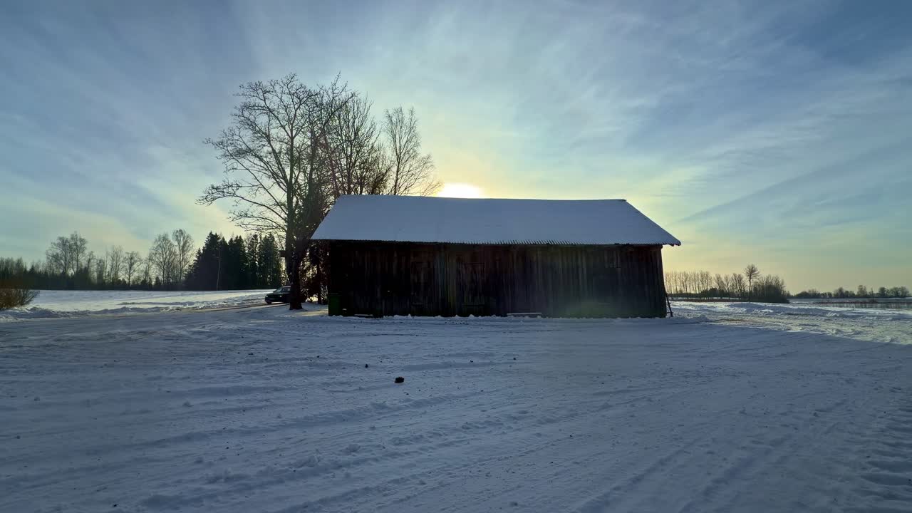 Bil, der sparker sne op og driver rundt på en frossen mark omkring en rustik lade ved solnedgang