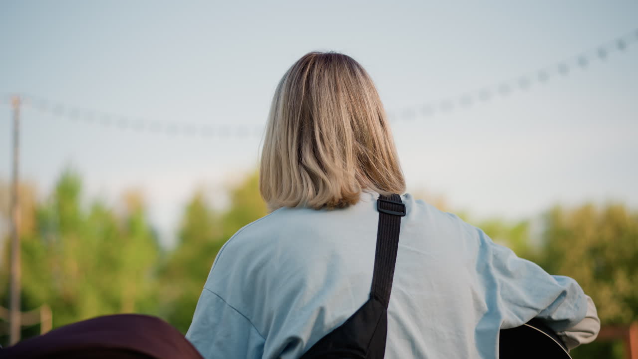 Blonde, blanke gitarist speelt akoestisch buiten bij zonsondergang, intiem profiel en achteraanzicht, casual spijkerjas, gitaarband over de schouder, oefent snaren en akkoorden, sfeervolle, beschouwende songwriter