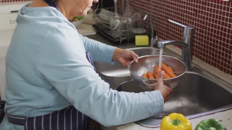 asian senior woman wearing an apron washing vegetables in the sink at home