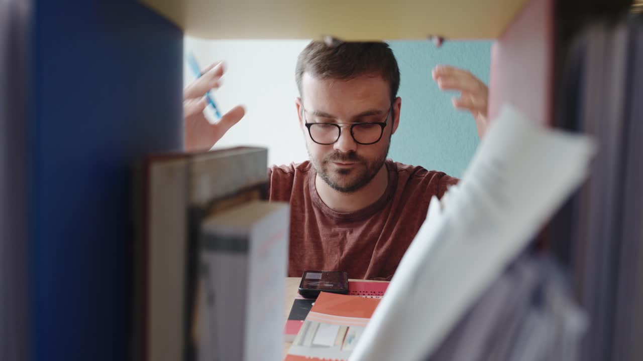 A young man browses through documents and binders on a shelf, clearly searching for something important. His expression shifts to disappointment as he fails to find what he was looking for
