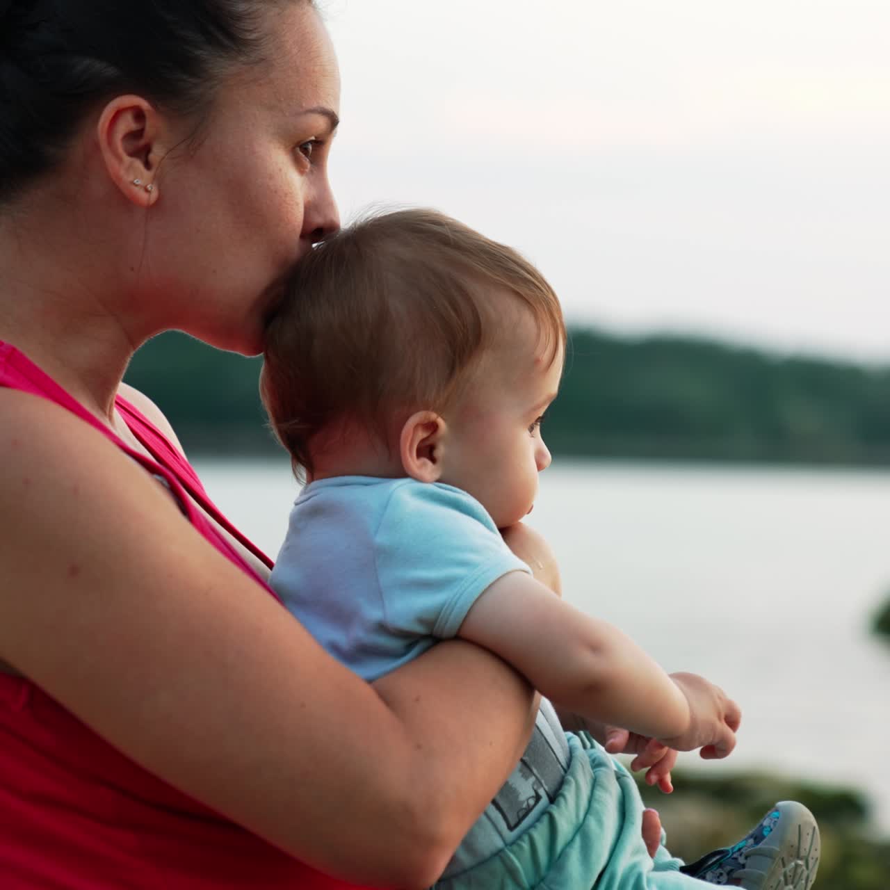 Mother is holding a baby in her arms sitting near the river in summer evening. Loving mommy kissing her son in head. Blurred backdrop