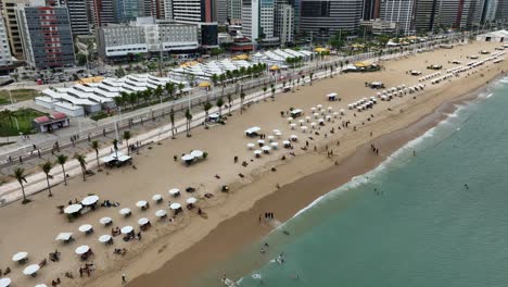 aerial flyover showing fortaleza beach with umbrellas, tourists, swimmers, and tall city buildings along the coastline