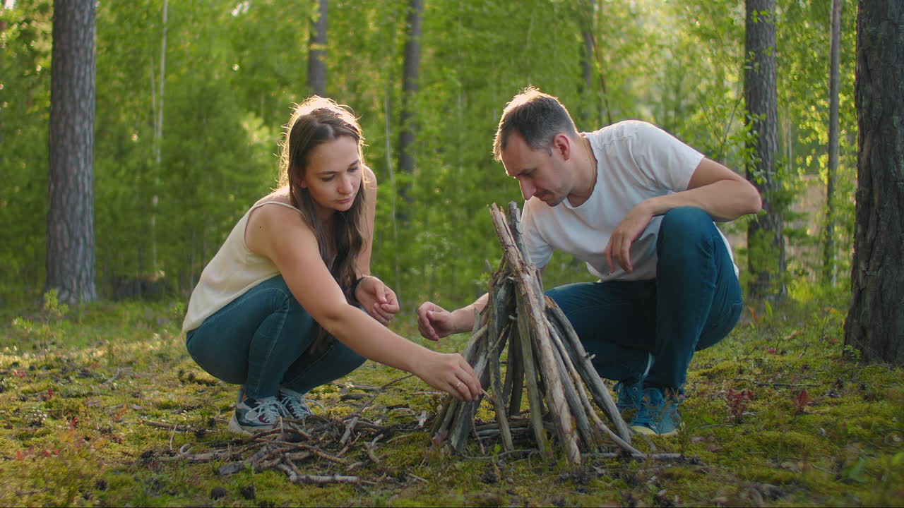 un hombre y una mujer juntos recogen un fuego de los palos en el bosque. una pareja joven en una caminata pone un fuego para la noche y cocinar en la naturaleza
