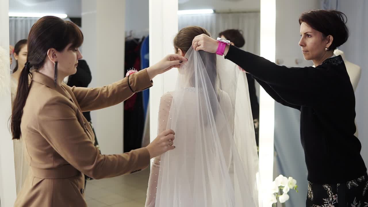 Rare view of happy bride trying her wedding gown in atelier, two female designers lifting her veil as she looks at her reflection in the mirror. Slow motion