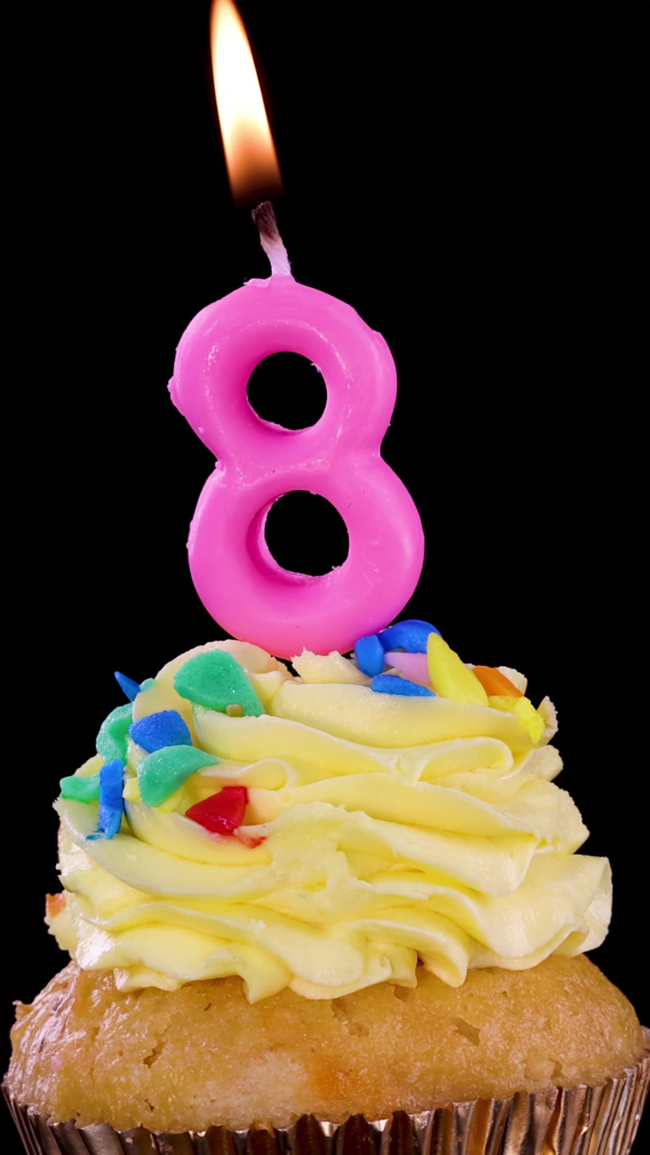 Candela rosa a forma di numero otto che brucia su un cupcake glassato colorato, con illuminazione drammatica e sfondo nero.