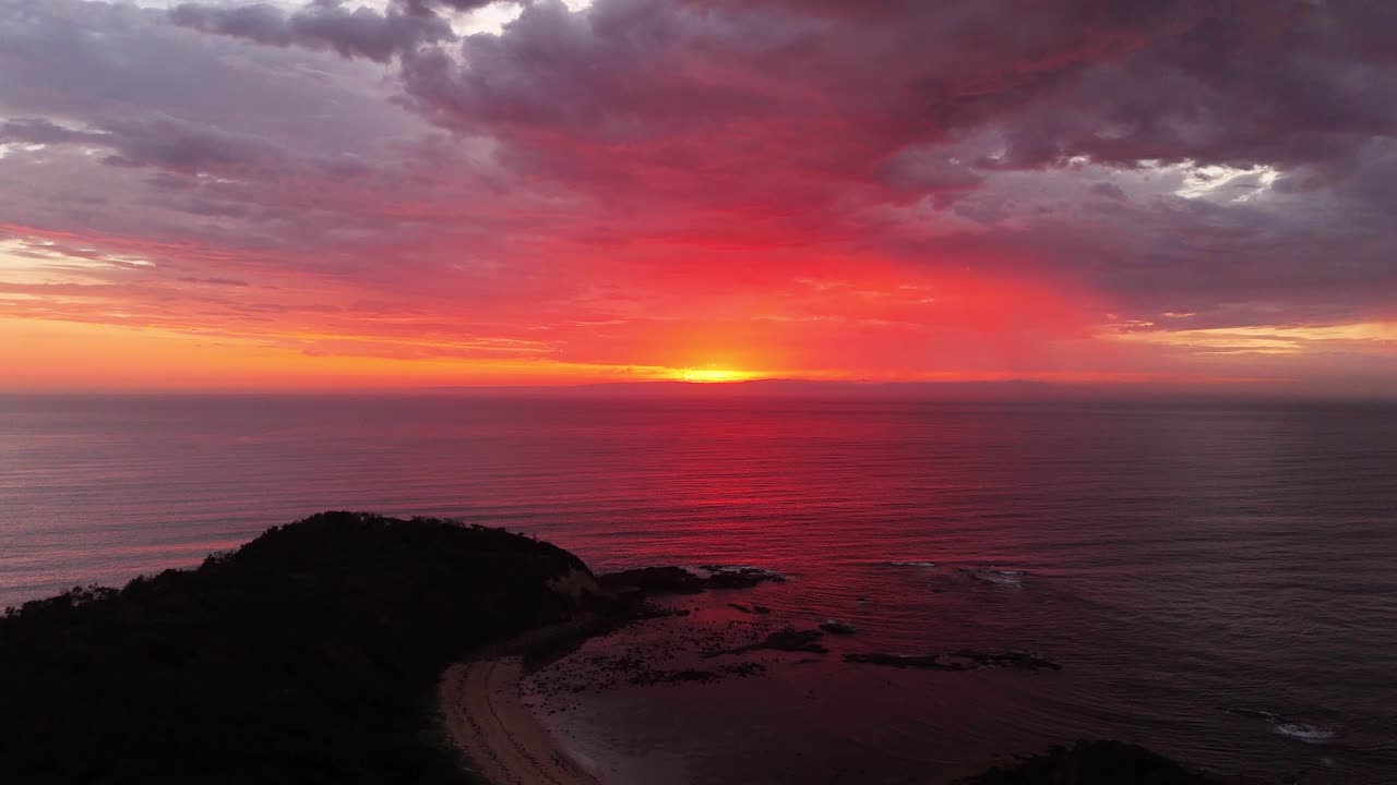 Aerial establishing pullback of a vibrant orange red sunrise over Inyadda Beach, NSW, Australia, with waves rolling onto the shore