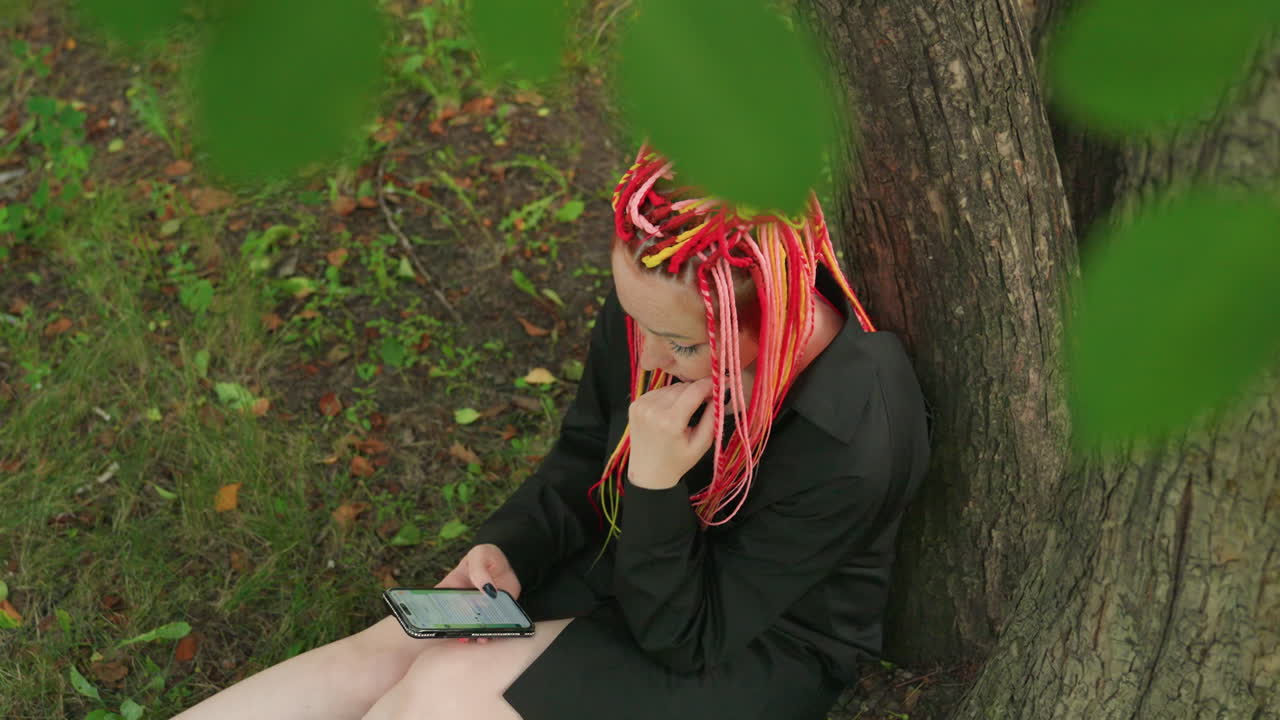 Белая женщина разговаривает по телефону под деревом, красные косы, крупный план профиля, вытирает слезу, виден экран смартфона, сидит на траве, интимное проявление эмоций, момент уединения в парке, выражение уязвимости