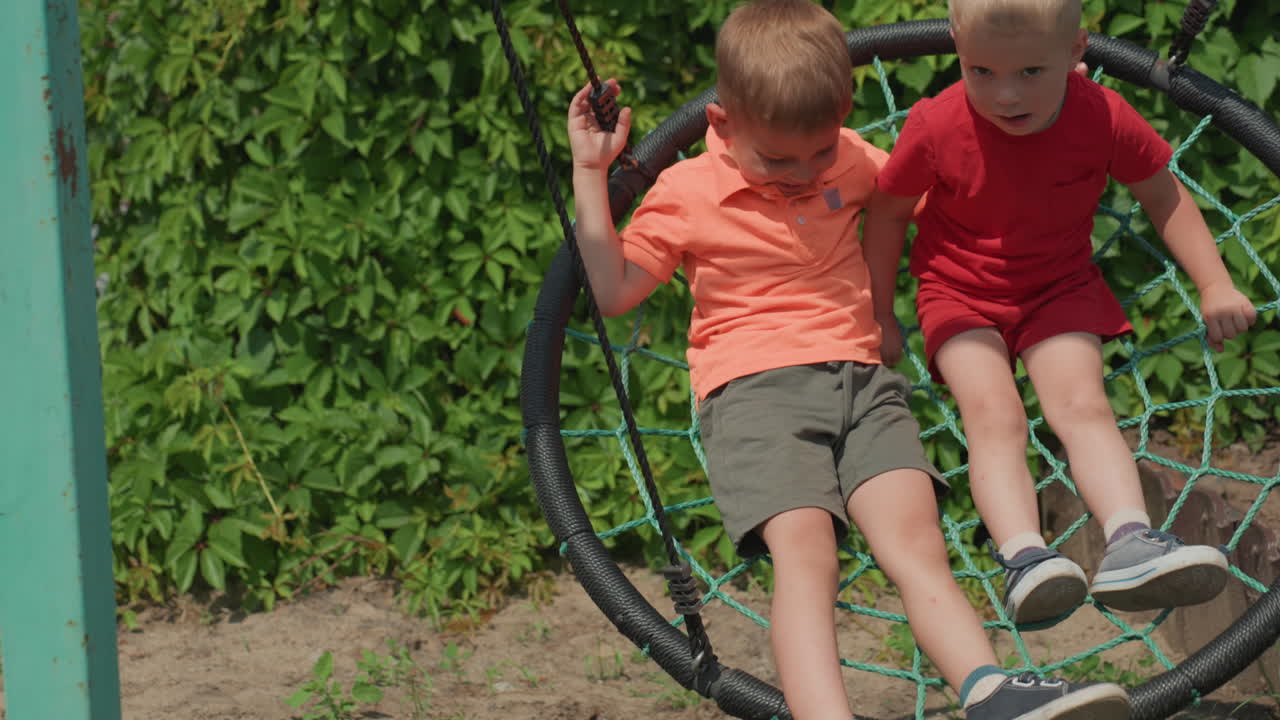 Caucasian Boy Smiling Into Camera Near Nest Swing In Backyard, Red Shirt And Curious Gaze, Friend Nearby And Leafy Fence Backdrop, Candid Summer Portrait Capturing Playful Innocence And Warm Light