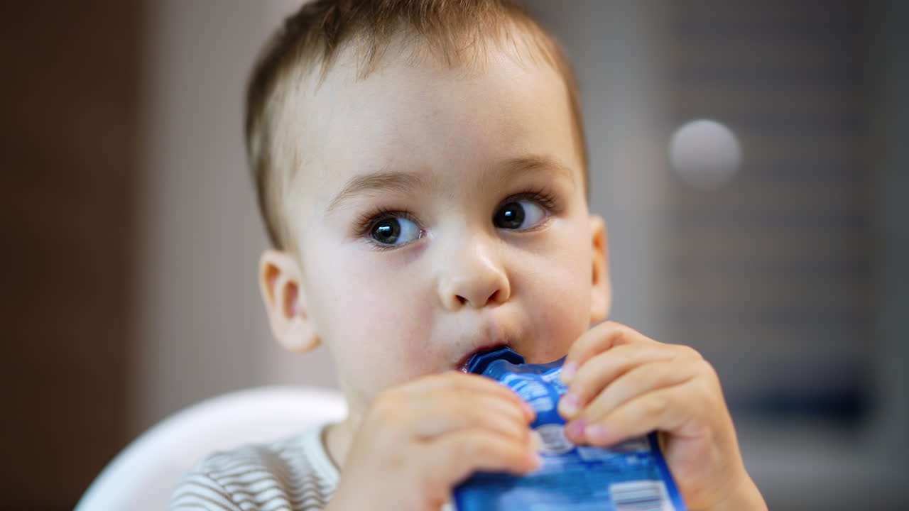Lovely Caucasian baby boy eating the fruit puree. Beautiful kid holding doy pack in both hands sucking the food. Close up.