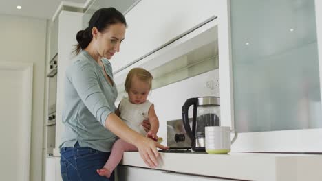 caucasian mother holding her baby using smartphone in the kitchen at home