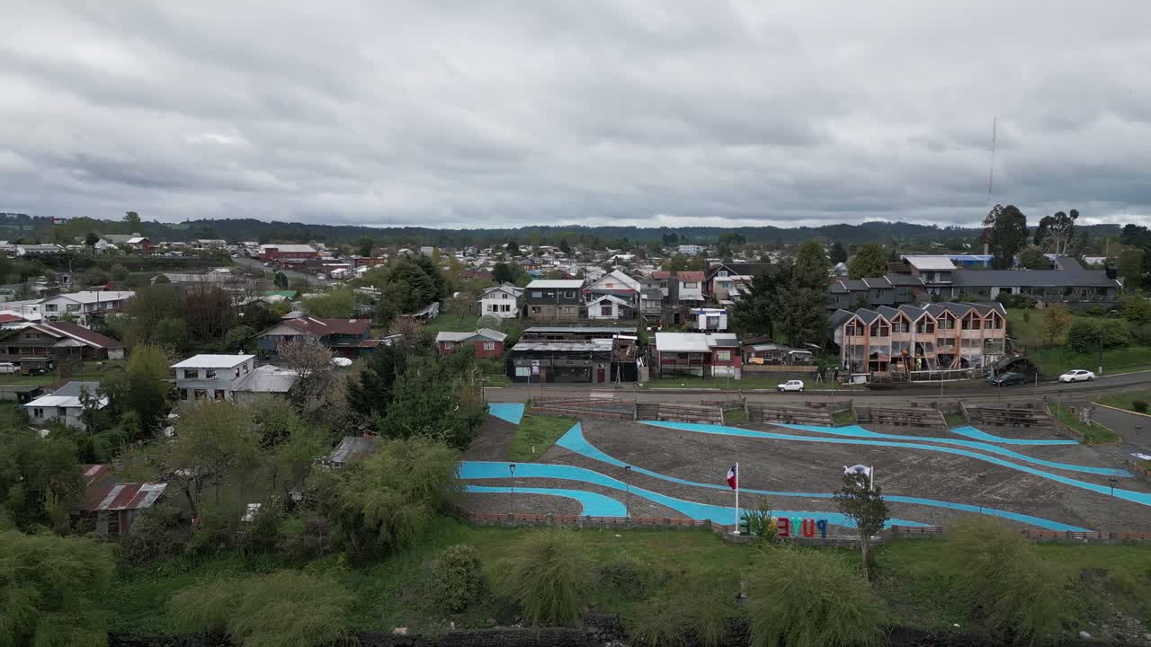 Panoraamailmakuva pienestä kaupungista, jossa on sinisiä katoksia aukiolla, ympäröitynä asuintaloilla ja vehreillä kukkuloilla dramaattisen pilvisen taivaan alla. Entre Lagos, Puyehue, Chile