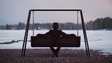 man sitting on a swing at the beach and looking towards the frozen lake