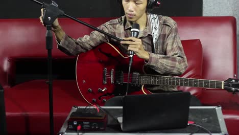 Medium-shot-songwriter-sit-on-red-sofa-adjusting-microphone-stand-positions-prepare-for-live-recording-in-black-and-white-studio