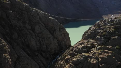 aerial shot flying through the valley between cliffs along the river out to the small, turquoise lake with a suspension bridge high up from side to side