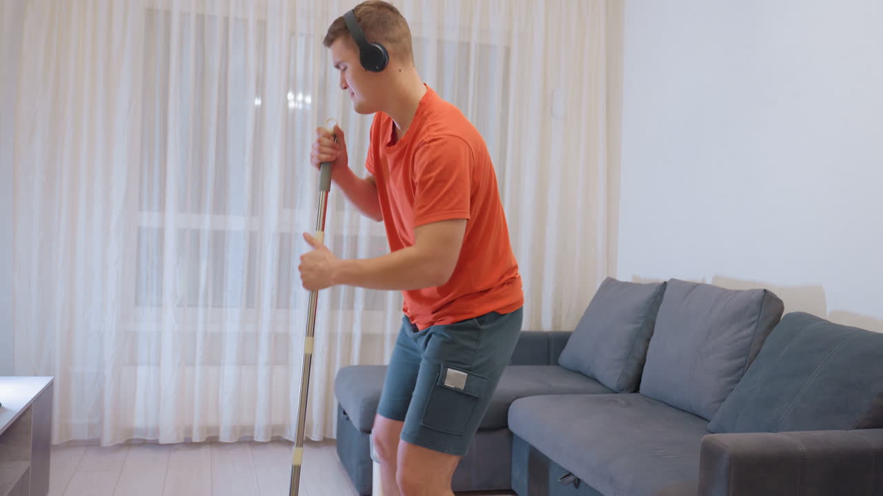 Cleaner in orange shirt and shorts dancing joyfully while holding mop on wooden floor near sofa enjoying music from headset turning household cleaning routine into fun