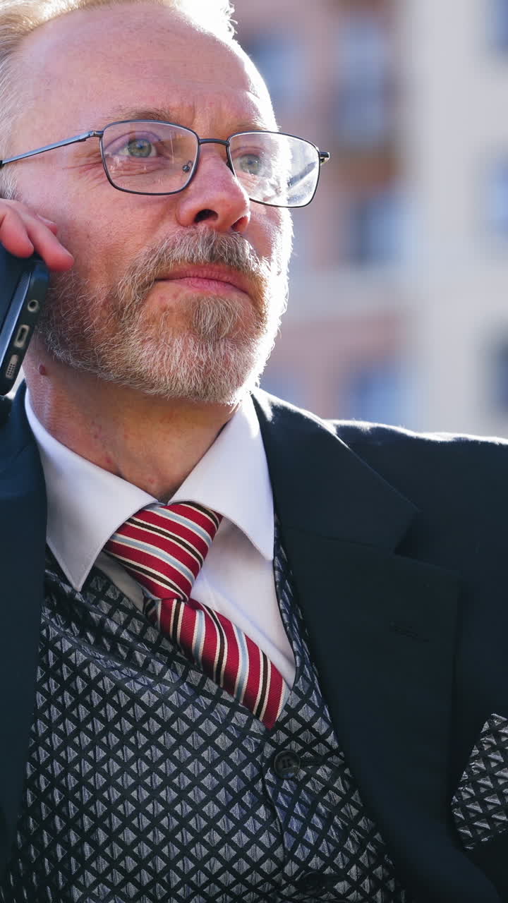 Happy elderly businessman talking on smartphone. Mature bearded man in suit and glasses sitting on bench and having a conversation about his business in the city. Vertical video