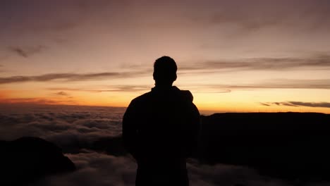 silhouetted man raises both arms in triumph above a sea of clouds during a vivid sunset on a mountain summit.