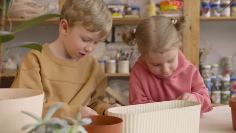 little blonde girl and blond kid preparing the soil in a pot sitting at a table where is plants in a craft workshop 2