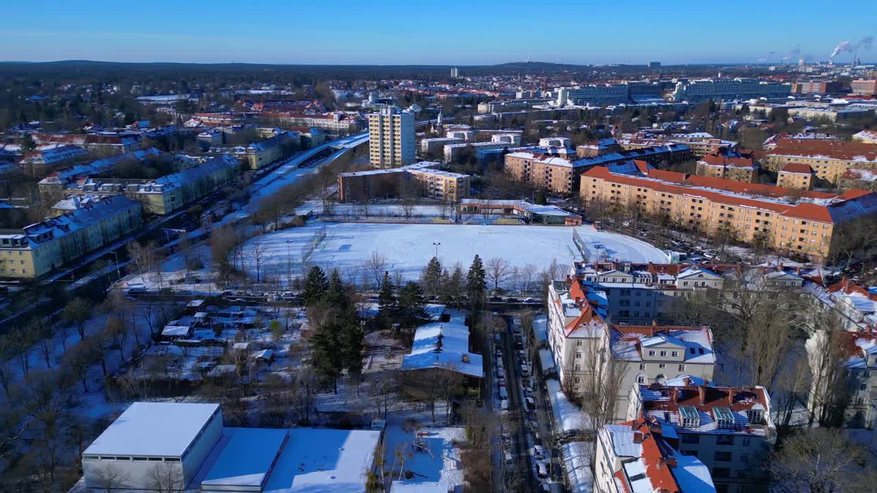 Berliinin kaupunkimaisema lumipeitteisellä jalkapallokentällä ja asuinrakennuksilla kylmänä talvipäivänä. Tasainen panoraama ilmakuva drone-kierrolla.