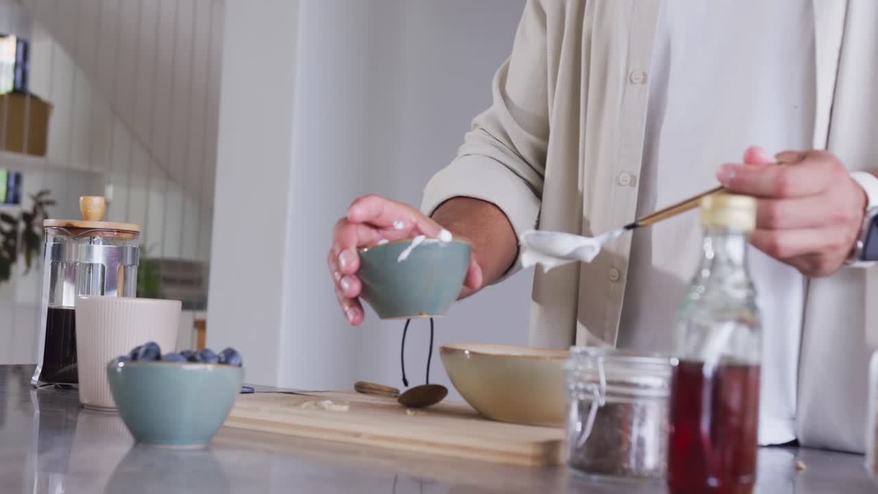 Uomo adulto che versa lo yogurt in una ciotola verde sull'isola della cucina, preparando la colazione con i mirtilli.