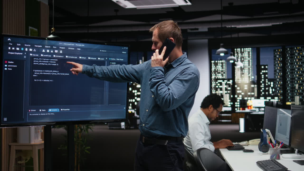 Software Developer Talking on Phone and Pointing at Code on Screen in Office
