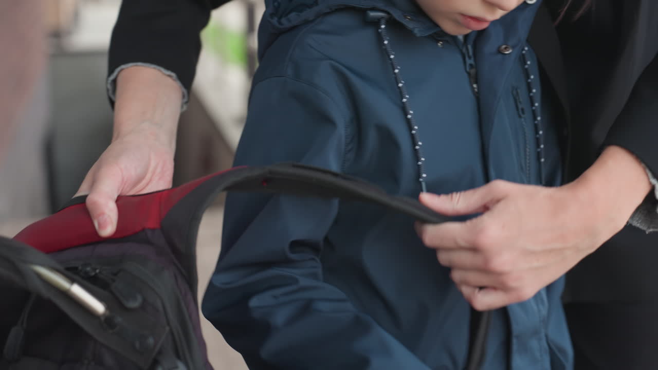 Niño blanco recibiendo una mochila de una mujer blanca, las manos ajustan las correas sobre los hombros, un toque firme, postura segura, detalle de la bolsa con forro rojo, acera matutina, preparación para la escuela, tierna despedida.