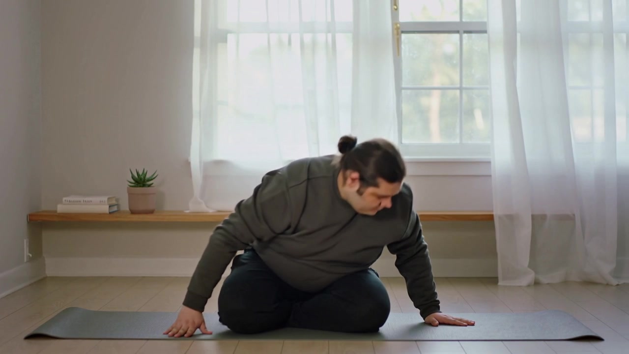 Seorang pria bersweater abu-abu mengangkat tangan, melakukan putaran ke kanan hingga berlutut di matras yoga untuk peregangan.