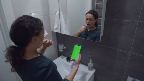 black woman looking at smartphone with chroma key while brushing teeth