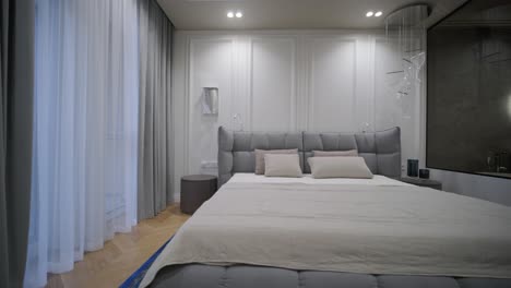 un dormitorio elegante con un cabecera de tufón gris, ropa de cama de lino beige y una paleta de colores neutrales, acentuada por la iluminación suave y las cortinas transparentes.